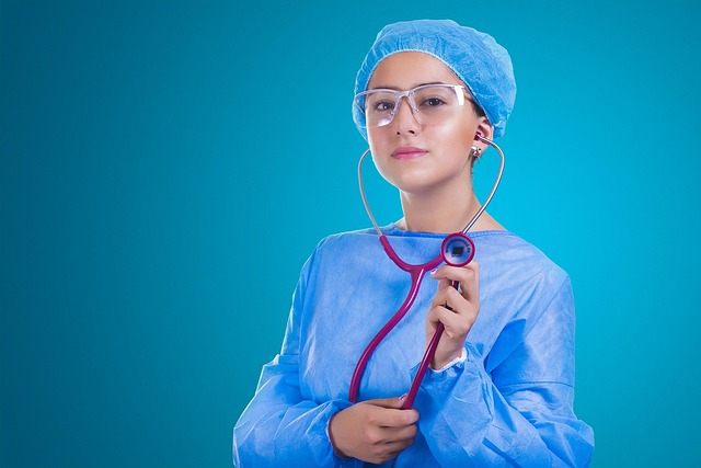 A medical professional wearing a blue surgical gown, cap, and safety glasses holds a stethoscope, standing against a blue background.