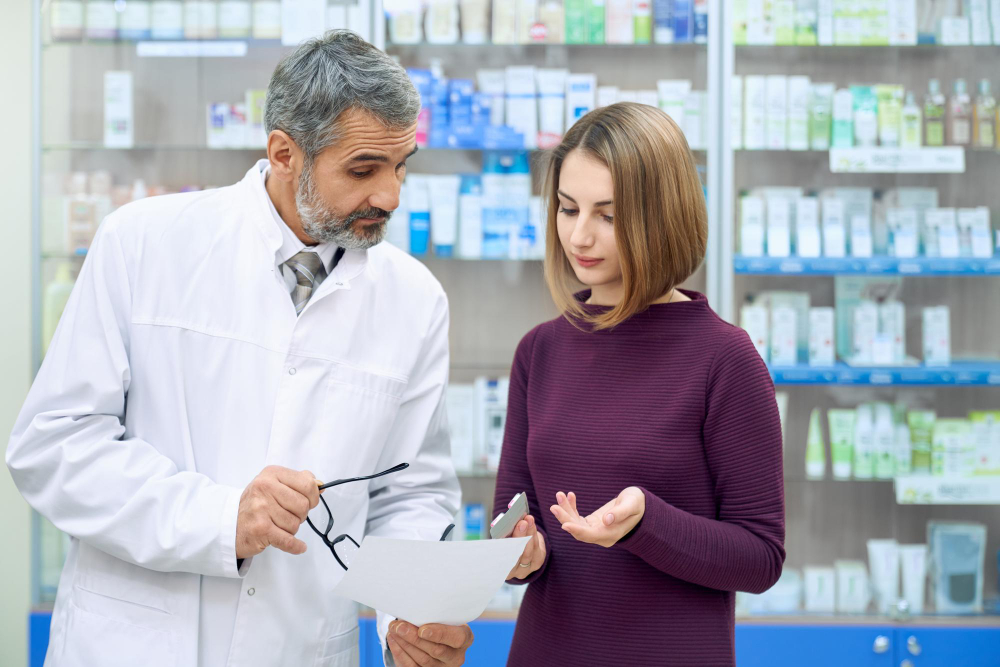 A pharmacist in a white coat explains a document to a woman in a pharmacy. The woman listens attentively, holding a phone, with shelves of medications and products visible in the background.