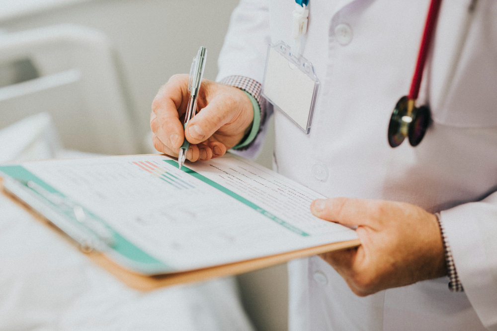 A doctor wearing a white coat and stethoscope fills out a medical form on a clipboard with a pen, focusing on documenting patient information.