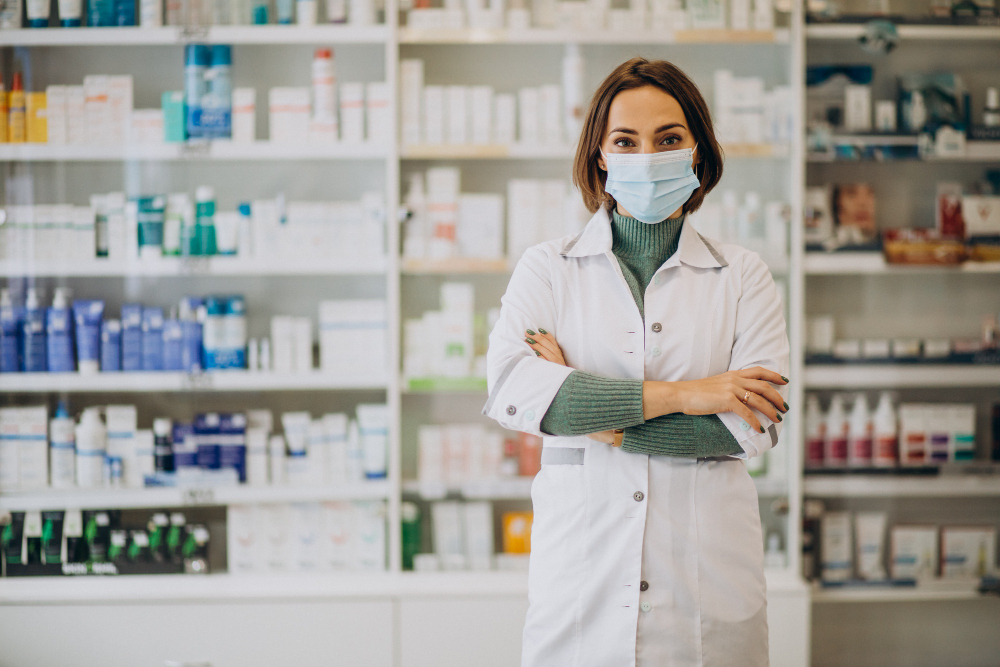 A pharmacist wearing a medical mask and white lab coat stands with arms crossed in front of shelves filled with medications and health products in a pharmacy.