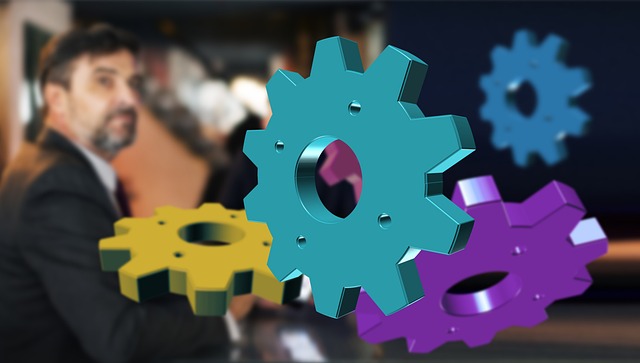 A blurred man in a suit sits in the background, while large, colorful gears (teal, yellow, and purple) are digitally superimposed in the foreground, symbolizing teamwork or business processes.