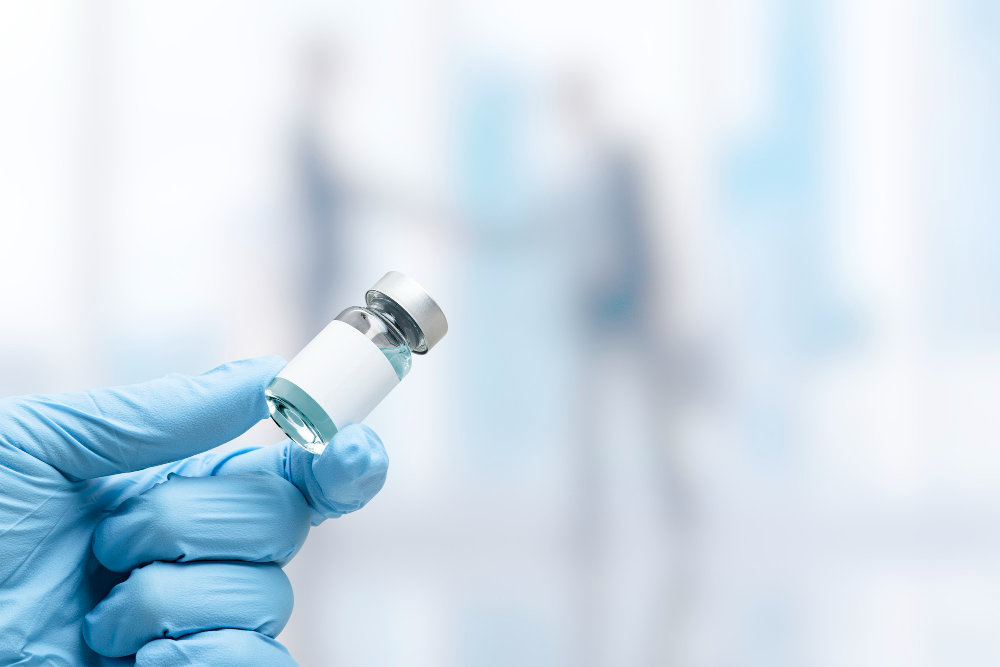 A gloved hand holds a small vial with a blank white label, containing clear liquid, against a blurred background showing two people shaking hands.
