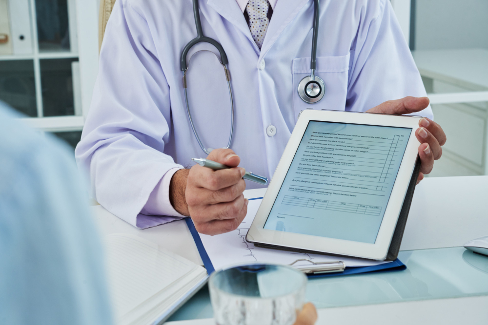 A doctor in a white coat shows a medical form on a tablet to a patient while holding a pen, suggesting a discussion about health information or treatment options during a consultation.